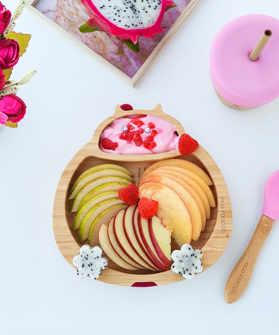 Eco Rascals Ladybird Bamboo Suction Plate filled with apple slices, pink yoghurt and decorated with fresh fruit, with a pink bamboo and silicone baby spoon and cup with a straw.