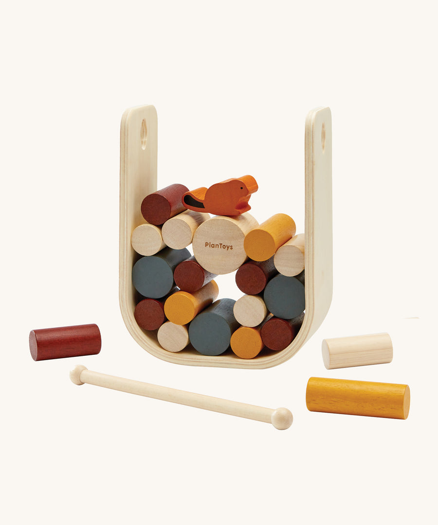 The PlanToys Giant Beaver Tumble is a fun kids game of strategic thinking and hand-eye coordination, using the wooden stick to push out the logs without the beaver falling. There is a large natural wood U shaped base to keep the sticks and beaver upright, and the sticks are in various sizes and colours such as yellow, brown, white and grey. The little beaver piece is orange with a black tal. The image is on a cream background.