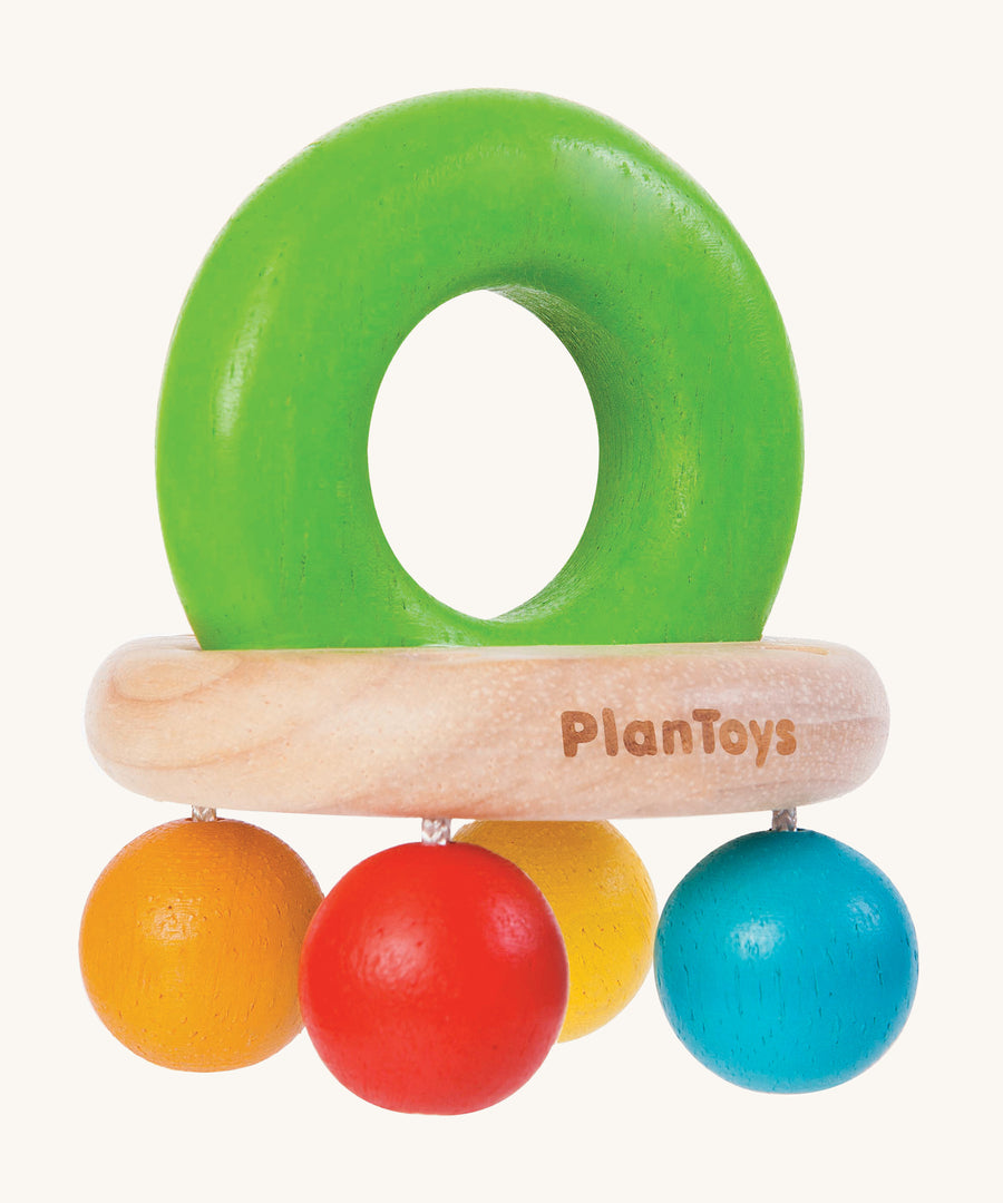 PlanToys Bell Rattle, is a beautiful wooden baby toy. The rattle comes in the shape of a bell, with a large green wooden ring on top for easy gripping. At the bottom of the ring is a horizontal wooden oval, and attached to the wooden oval are four bright coloured balls in blue, orange, red and yellow. The image is on a cream background