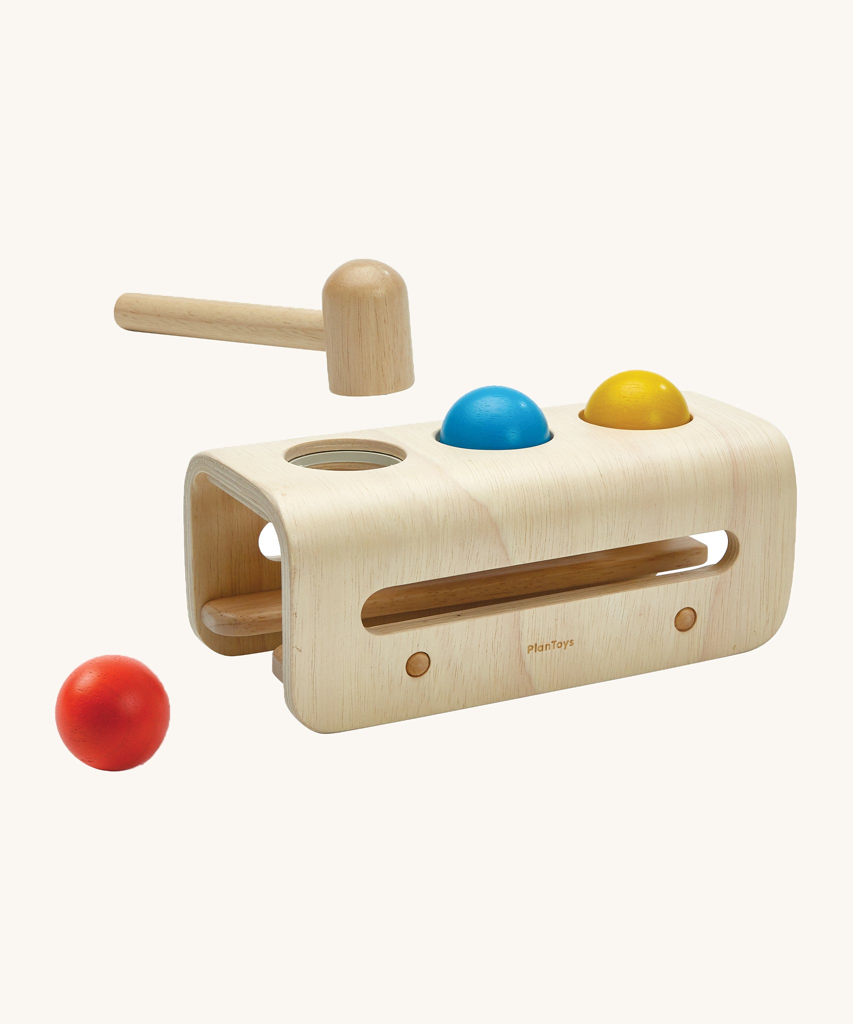 The PlanToys wooden Hammer Ball play set has a rounded base to allow for movement when the balls are being hammered through the holes. There are two wooden horizontal sticks in the underside of the hammer bench to allow the balls to run out and be collected. The balls are red, blue and yellow and there is a wooden hammer/mallet above the hammer board.