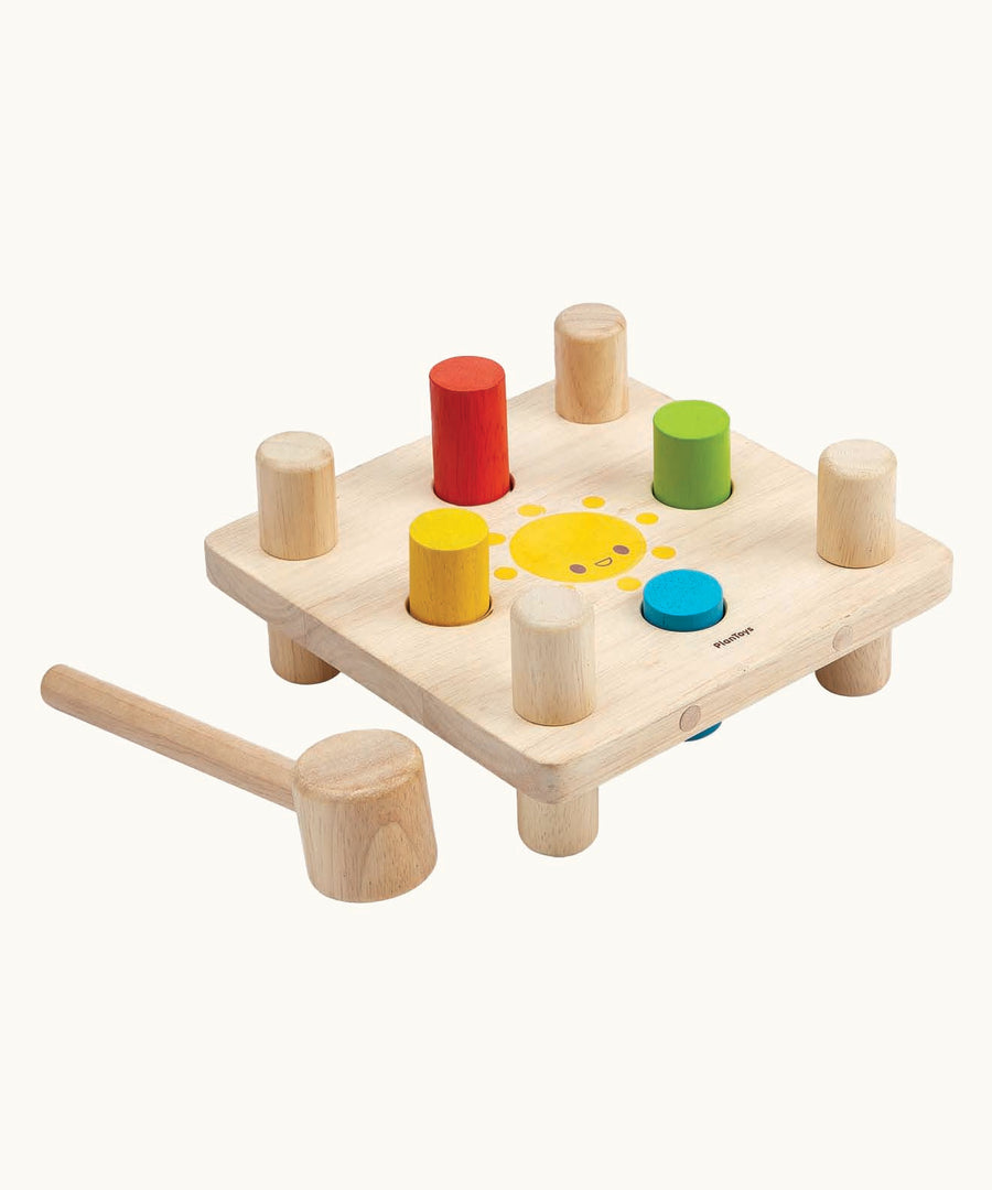 The Plan Toys Wooden Hammer Peg  toy, is a classic wooden toy hammer bench with colourful pegs for babies and toddlers. The bench is square in shape with a smiling sun in the middle, and has a red, yellow, blue and green peg. There is also a wooden hammer next to the peg board. The image is on a cream background.