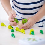 A child placing the green cactus pegs into the natural wooden block. The block has two cactus pieces balanced, with the other green cactus pieces scattered on a light coloured table just below the child's hands