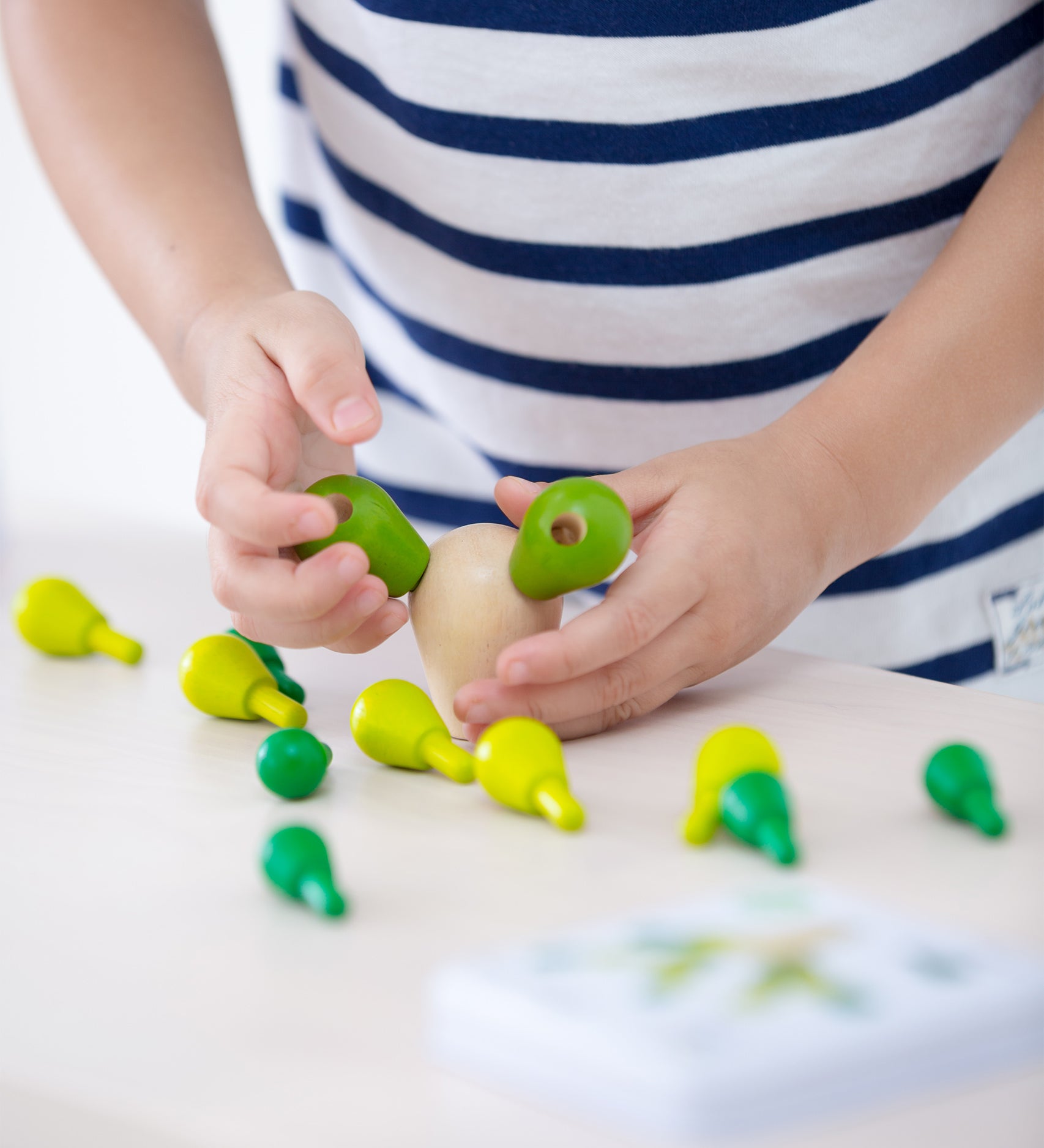 A child placing the green cactus pegs into the natural wooden block. The block has two cactus pieces balanced, with the other green cactus pieces scattered on a light coloured table just below the child's hands