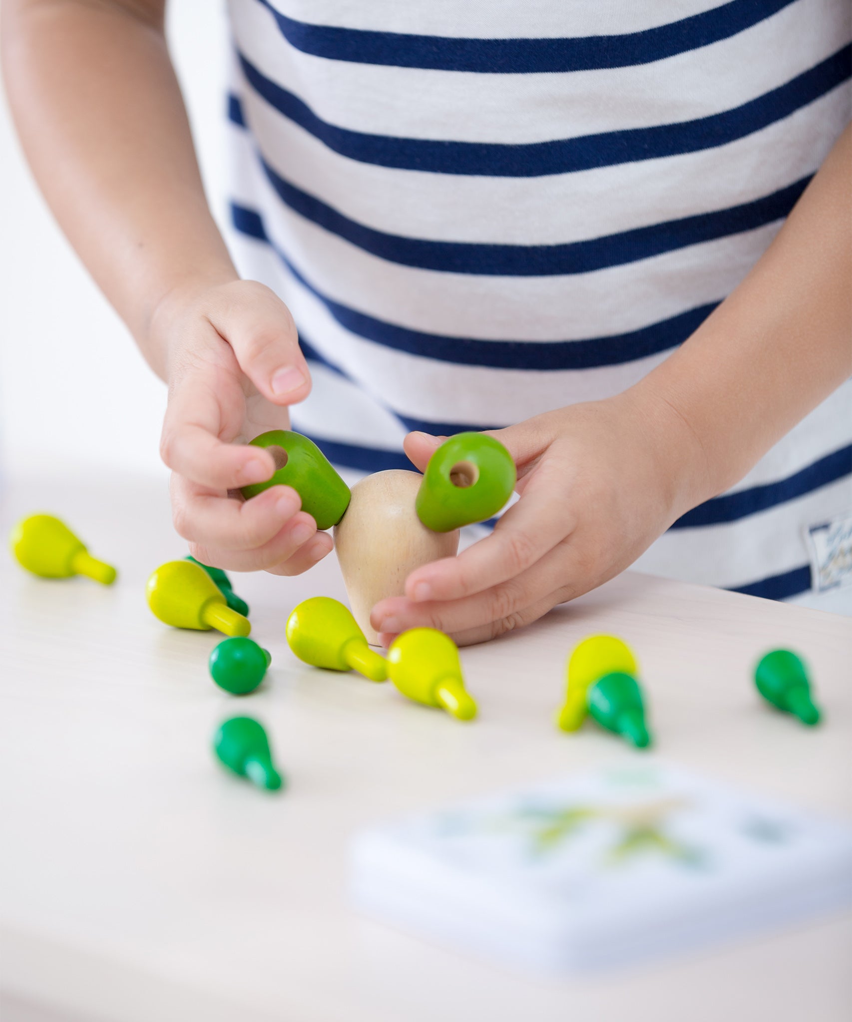 A child placing the green cactus pegs into the natural wooden block. The block has two cactus pieces balanced, with the other green cactus pieces scattered on a light coloured table just below the child's hands