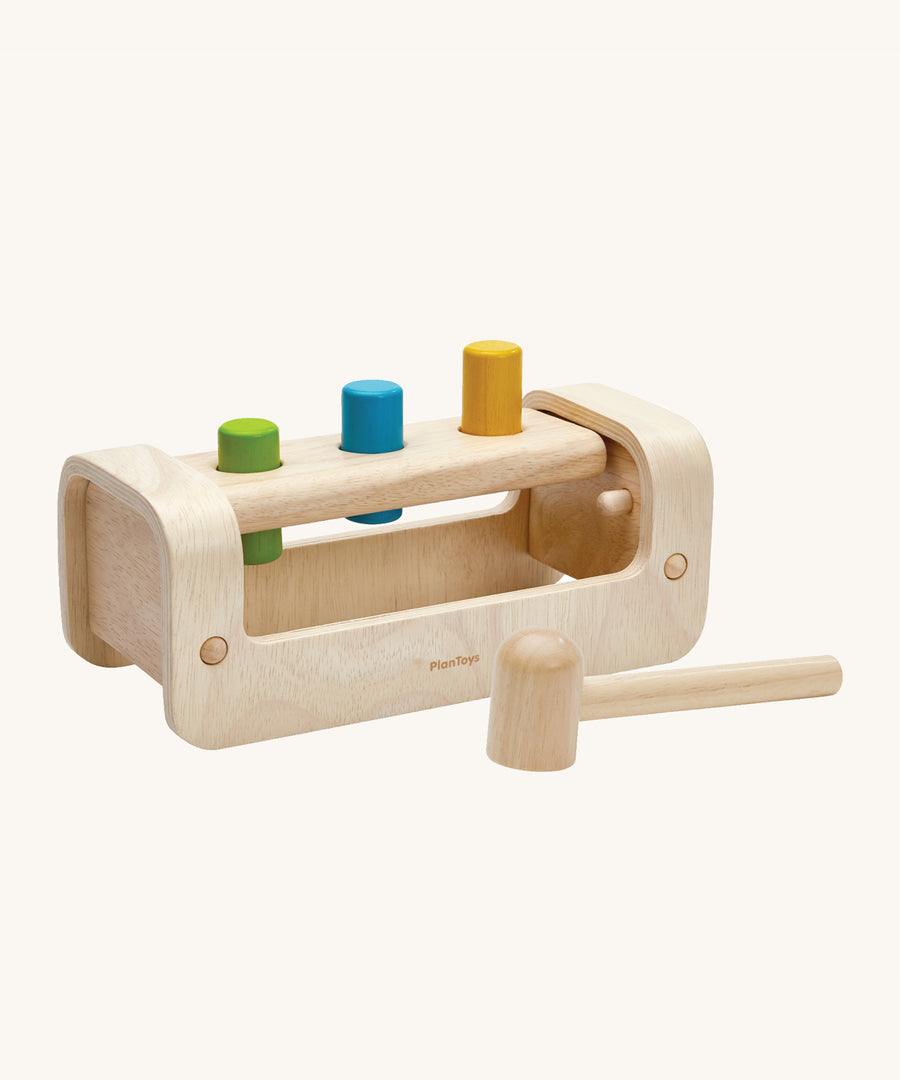 The hammer/mallet is in front of the PlanToys Pounding Bench. The hammer/mallet has one flat side and one rounded side. The image is on a cream background