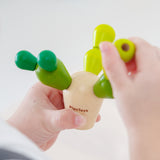 A child placing the green cactus pegs into the natural wooden cactus block to see if the pieces balance or topple over. The child is holding the toy in their hands