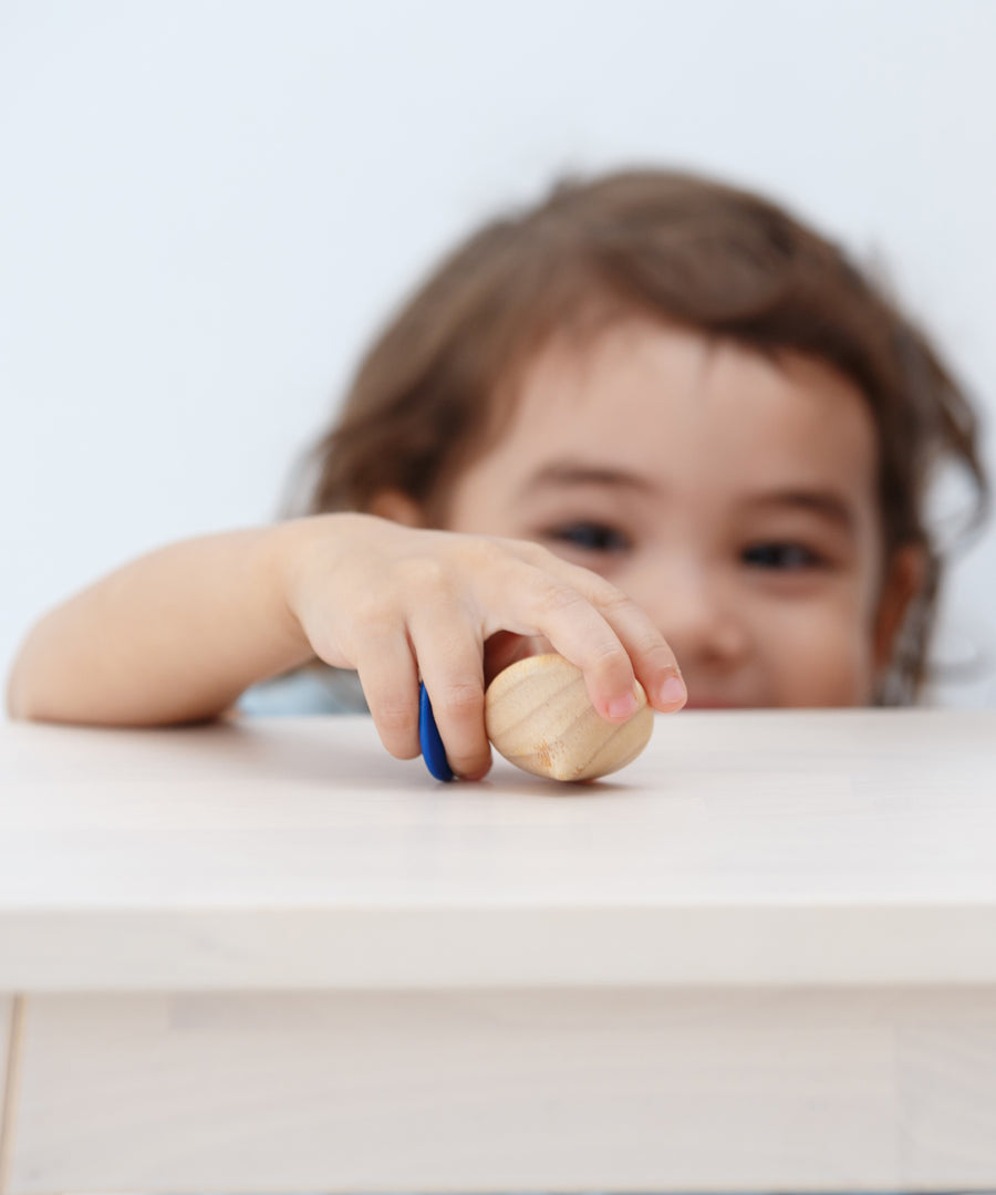 A child holding the PlanToys Mini Spinning Tops in their hand on a wooden table top