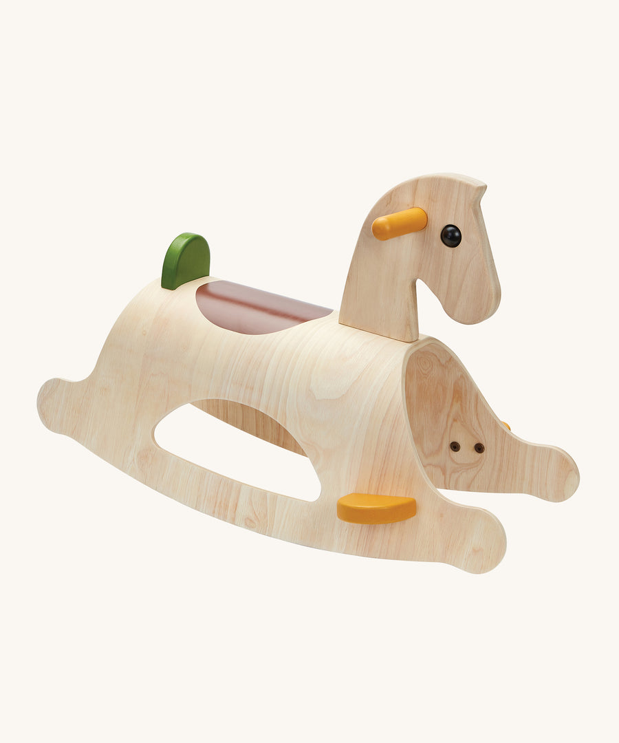 PlanToys Palomino Rocking Horse - Modern Rustic is a beautiful first rocking horse for any child. This rocking horse is in natural wood showing the wood-grain, with yellow handles and foot plates, a brown seat patch on the back of the horse, and a green back rest. The image is on a cream background