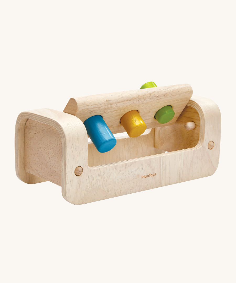 The peg board lifted up on the PlanToys Pounding Bench, to show the pegs going through the holes. Little wooden dowels allow the peg board to stay in place when hammering, and to move upwards to collect the pegs. The image is on a cream background