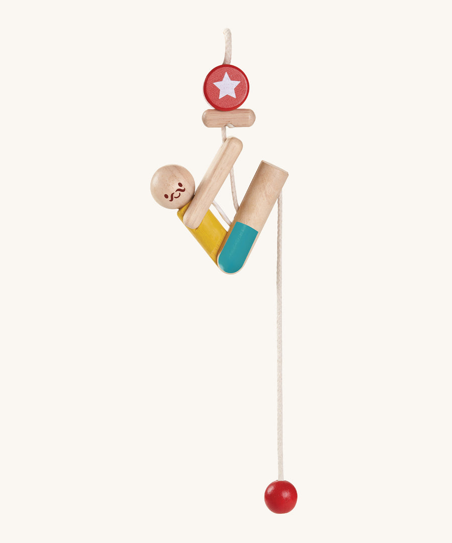 The PlanToys Rope Climbing Acrobat is a fun little wooden figure, on a rope. The acrobat is made fron natural wood pieces, and has a yellow painted top and blue painted shorts. There s a red circle with a star at the top of the rope, and a red ball at the bottom. The image is on a cream background