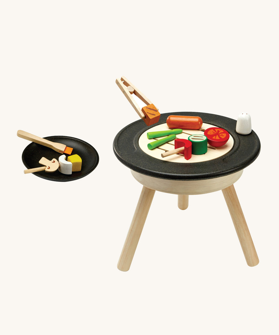 PlanToys kids wooden BBQ role play set which includes a selection of BBQ play food, skewers, basting brush, salt shaker, plate and a standing BBQ. The image is on a cream background