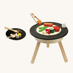 PlanToys kids wooden BBQ role play set which includes a selection of BBQ play food, skewers, basting brush, salt shaker, plate and a standing BBQ. The image is on a cream background