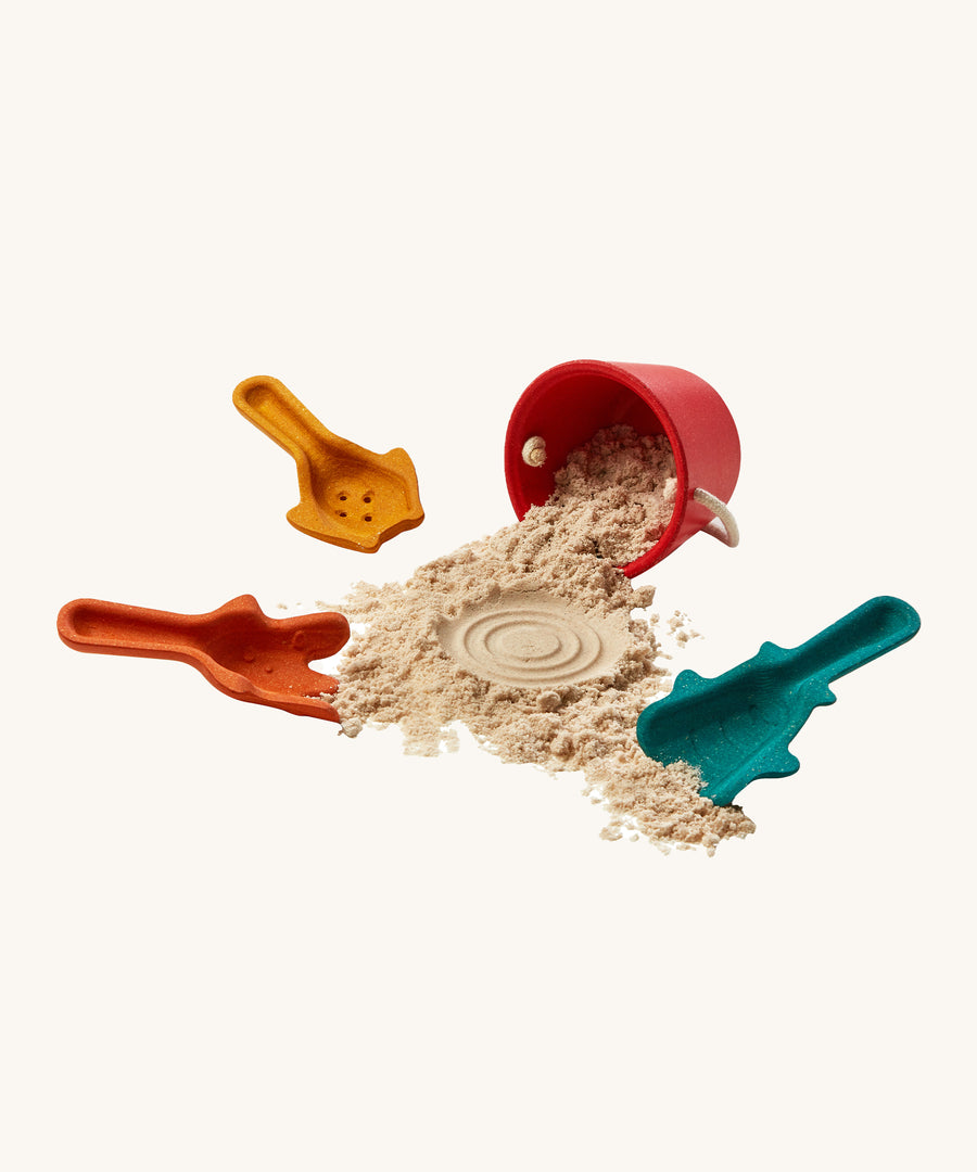 The PlanToys Sand Play bucket with sand inside, and scoops on a cream background