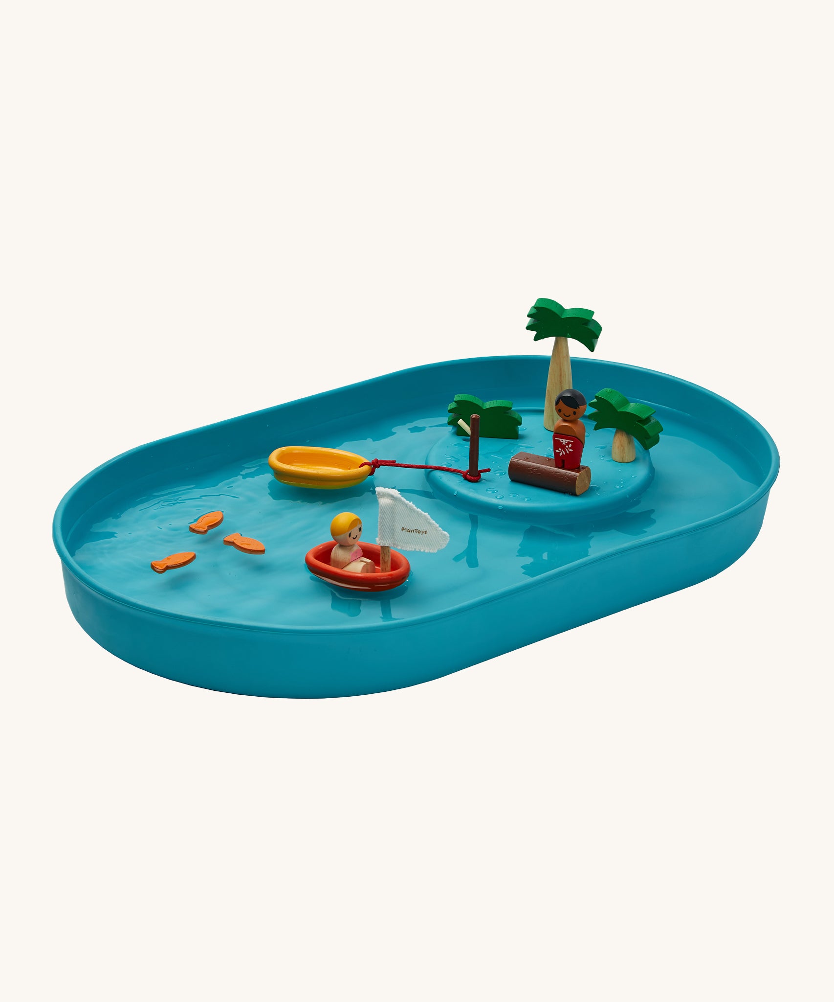 PlanToys Water Play Set is made from natural rubber and is great for sensory water play. The image shows a blue tray filled with water, with 3 fish floating on the water, a figure in a PlanToys boat, a boat tied to a brown pole and a figure stood on a wood log with palm trees to give a raised island effect. The image is on a cream background