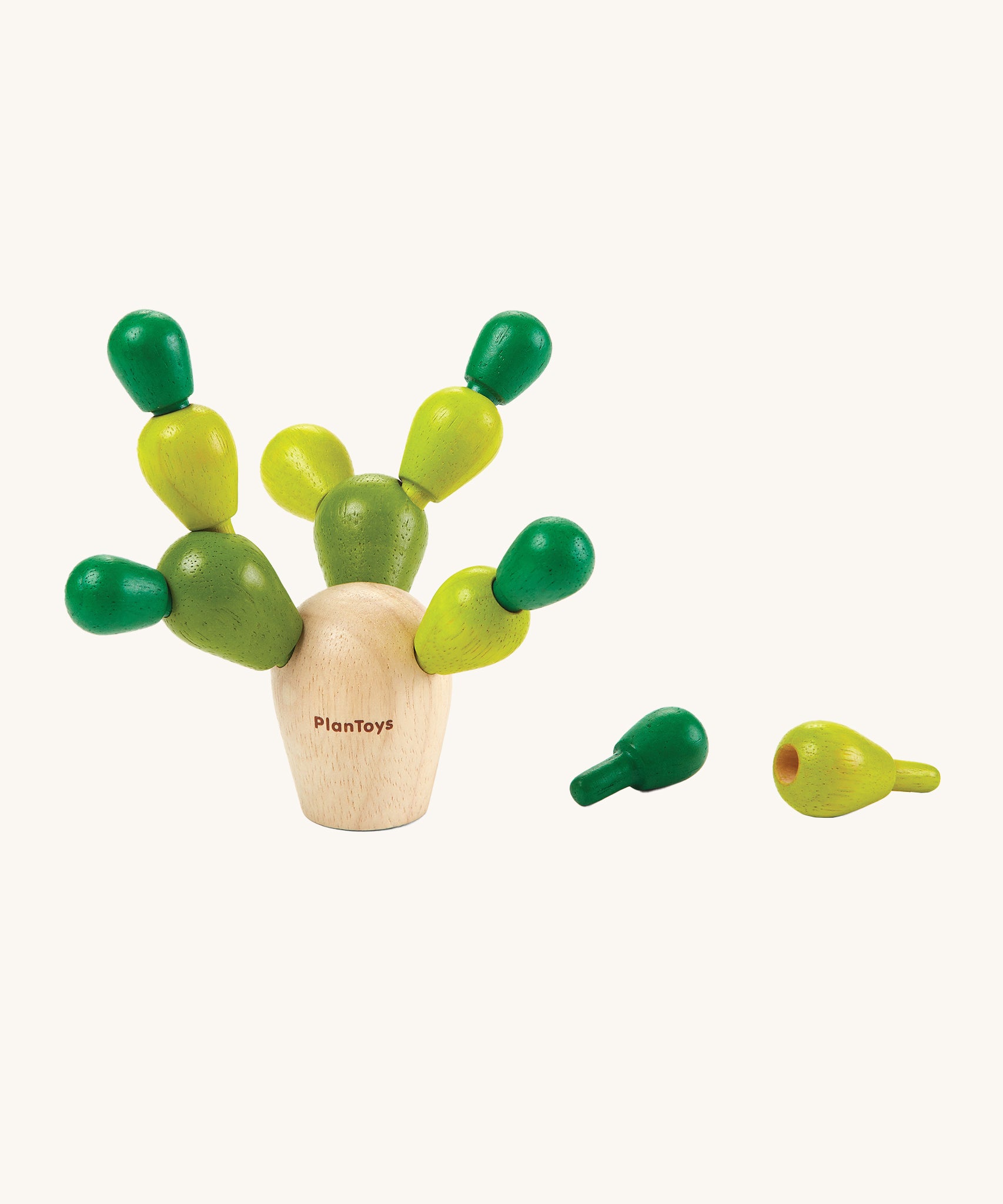 The PlanToys Mini Balancing Cactus with the green cactus pegs balanced carefully in the natural wooden cactus block, with two green cactus pegs to the right side of the cactus, on a cream background