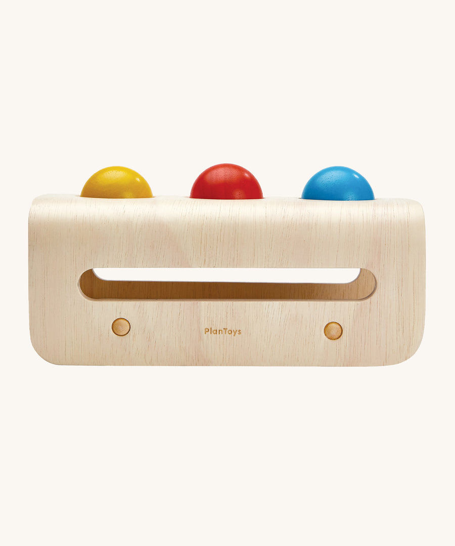 A side view of the PlanToys Hammer Balls set showing the yellow, rd, and blue ball resting on top of the hammer board. The image is on a cream background