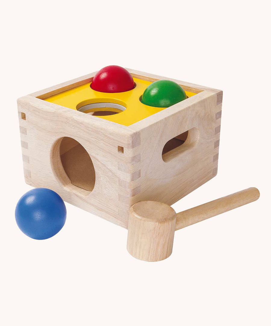 This PlanToys Punch and Drop game is a classic wooden pounding toy that babies and toddlers love – use the hammer to gently whack the three coloured balls through the holes in the wooden box and watch as they roll out. The ball colours are blue, red and green, and the top of the pounding box is yellow. The image is on a cream background