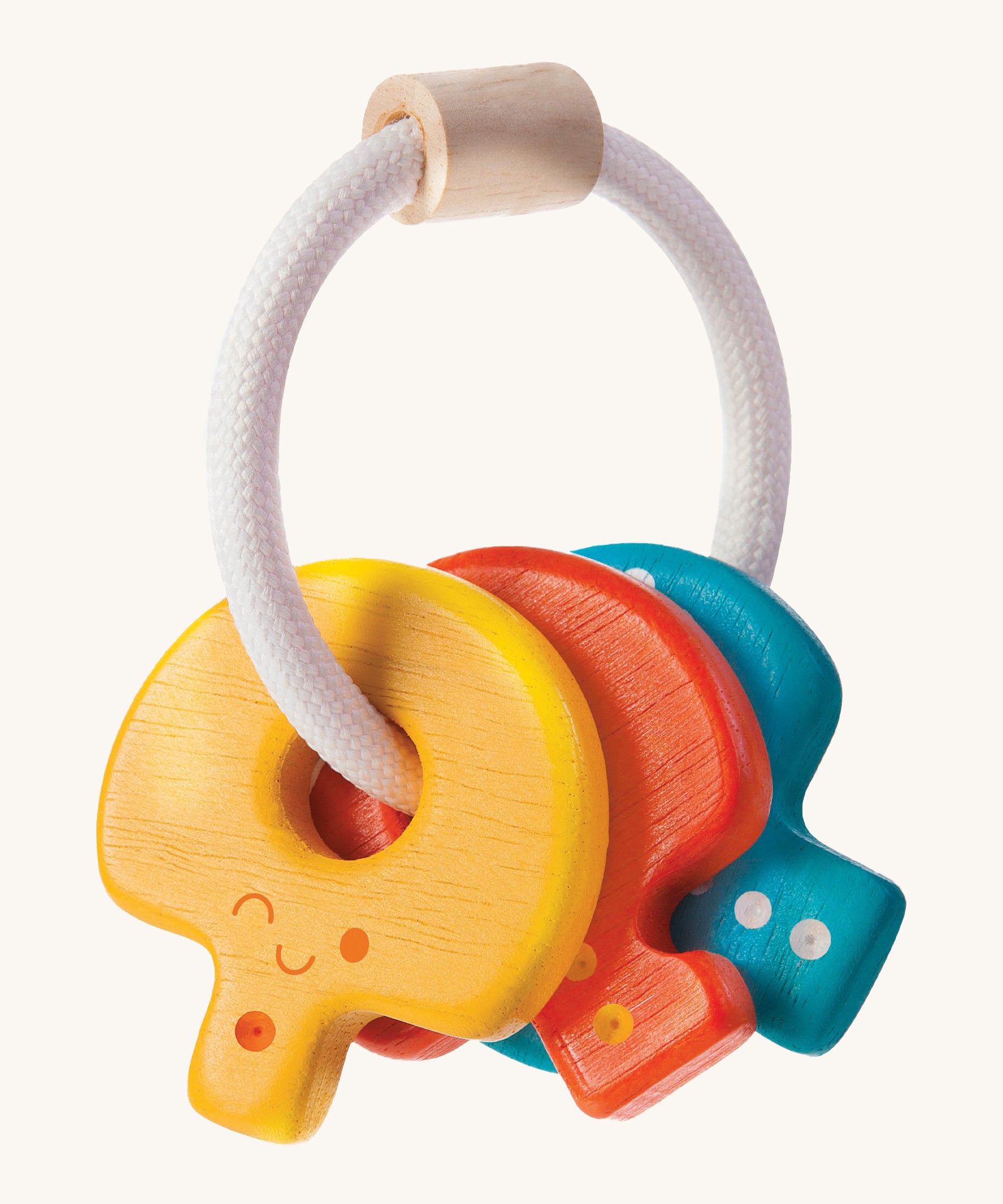 PlanToys Baby Key Rattle is a fun sensory toy for little ones. This toy is great for teething and soothing sore gums. There is a circular white rope with a wooden bead at the top. Attached to the rope are three, colourful wooden keys, all with smiling faces and dots to correspond to the key. The yellow key has one dot, the red key has two dots, and the blue key has three dots. The image is on a cream background.