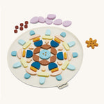 The PlanToys Mandala Set, is a beautiful wooden set of shapes in various colours. In the image, the shapes are laid out in a symmetrical pattern on a which circular mat, with dots, petals, and a flower shape at he top of the mat. The image is on a cream background