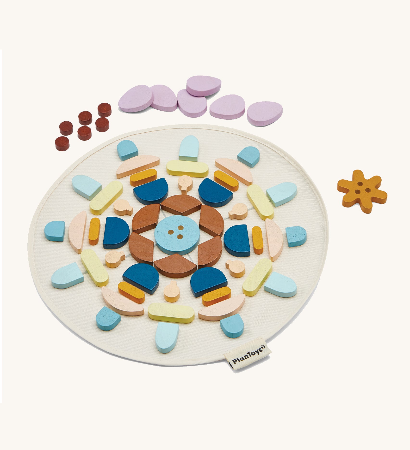 The PlanToys Mandala Set, is a beautiful wooden set of shapes in various colours. In the image, the shapes are laid out in a symmetrical pattern on a which circular mat, with dots, petals, and a flower shape at he top of the mat. The image is on a cream background