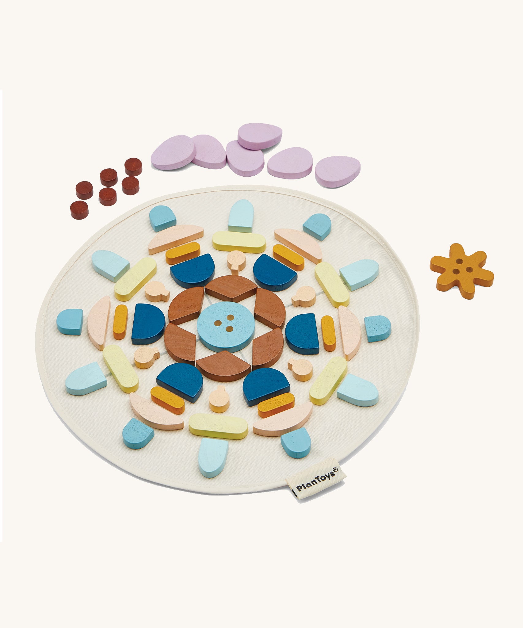 The PlanToys Mandala Set, is a beautiful wooden set of shapes in various colours. In the image, the shapes are laid out in a symmetrical pattern on a which circular mat, with dots, petals, and a flower shape at he top of the mat. The image is on a cream background