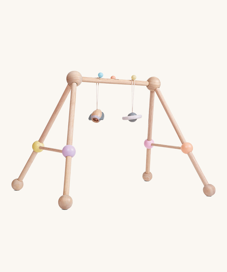 The PlanToys Play Gym Orchard is a fun, sensory baby gym made from wood. This set includes a rocket and a planet toy attached to the top of the baby gym using a pastel blue and a yellow peg. This is a side view image on a cream background.