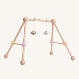 The PlanToys Play Gym Orchard is a fun, sensory baby gym made from wood. This set includes a rocket and a planet toy attached to the top of the baby gym using a pastel blue and a yellow peg. This is a side view image on a cream background.