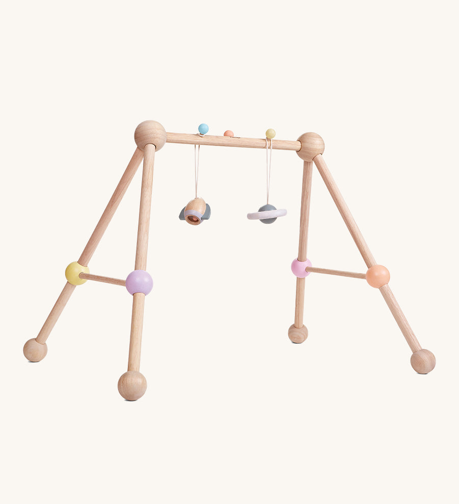 The PlanToys Play Gym Orchard is a fun, sensory baby gym made from wood. This set includes a rocket and a planet toy attached to the top of the baby gym using a pastel blue and a yellow peg. This is a side view image on a cream background.