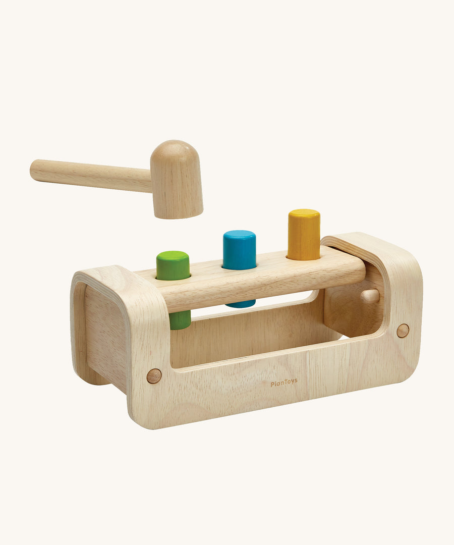 PlanToys Wooden Pounding Bench, has a curved, semi-circular base with a rectangle peg board with 3 holes, to allow for 3 wooden pegs to go through. The base allows for movement of the toy in a rocking motion. In this image there is a wooden hammer/mallet above the pegs in the peg board and the peg colours are green, blue and yellow. This image is on a cream background