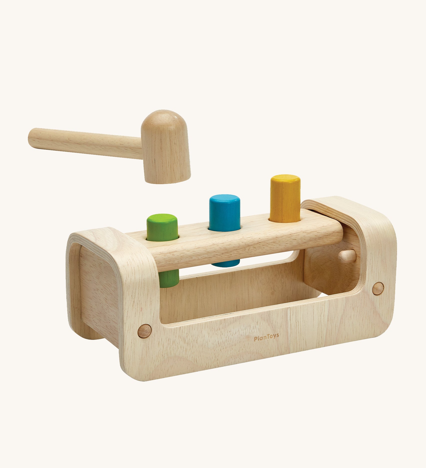 PlanToys Wooden Pounding Bench, has a curved, semi-circular base with a rectangle peg board with 3 holes, to allow for 3 wooden pegs to go through. The base allows for movement of the toy in a rocking motion. In this image there is a wooden hammer/mallet above the pegs in the peg board and the peg colours are green, blue and yellow. This image is on a cream background