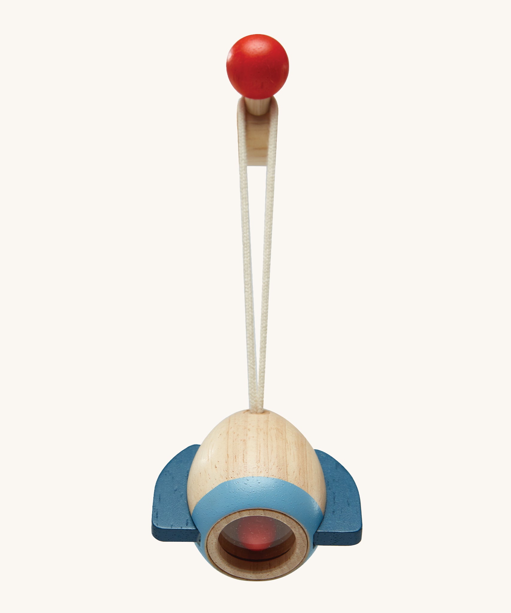 A closer look at the rocket toy from the PlanToys Play Gym in Orchard colours, on a red ball hook. The rocket is made from natural wood showing the wood grain and a light blue band at the bottom, dark blue rocket fins, and there is also a red ball inside. The image is on a cream background