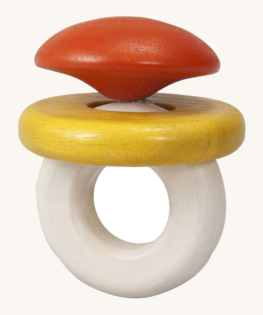 This PlanToys Baby Rattle, is in the shape of a ring, with an orange/red diamond jewel shape on top, a white ring attached for easy grasping, and a horizontal yellow ring which moves. The image is on a cream background