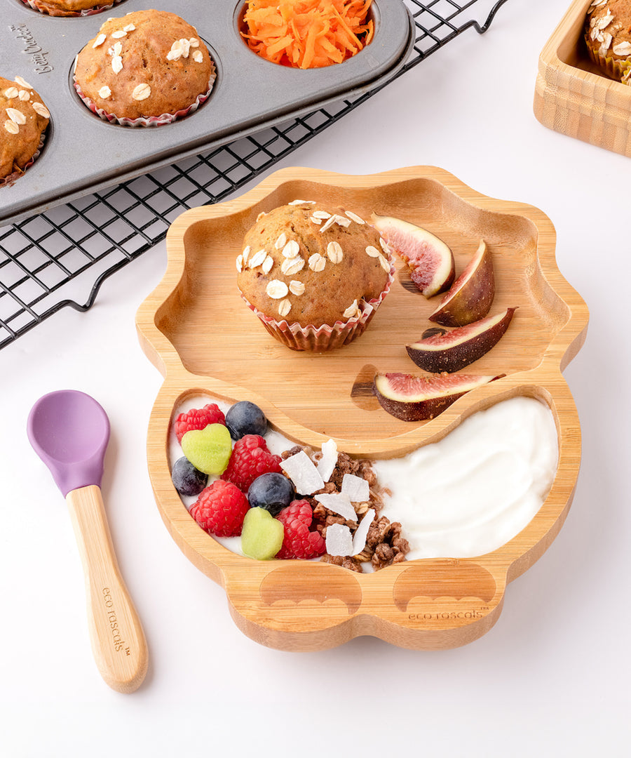 Eco Rascals Dinosaur Bamboo Suction Plate with a muffin, fig slices, yogurt and fresh fruit, and purple silicone and bamboo baby spoon.