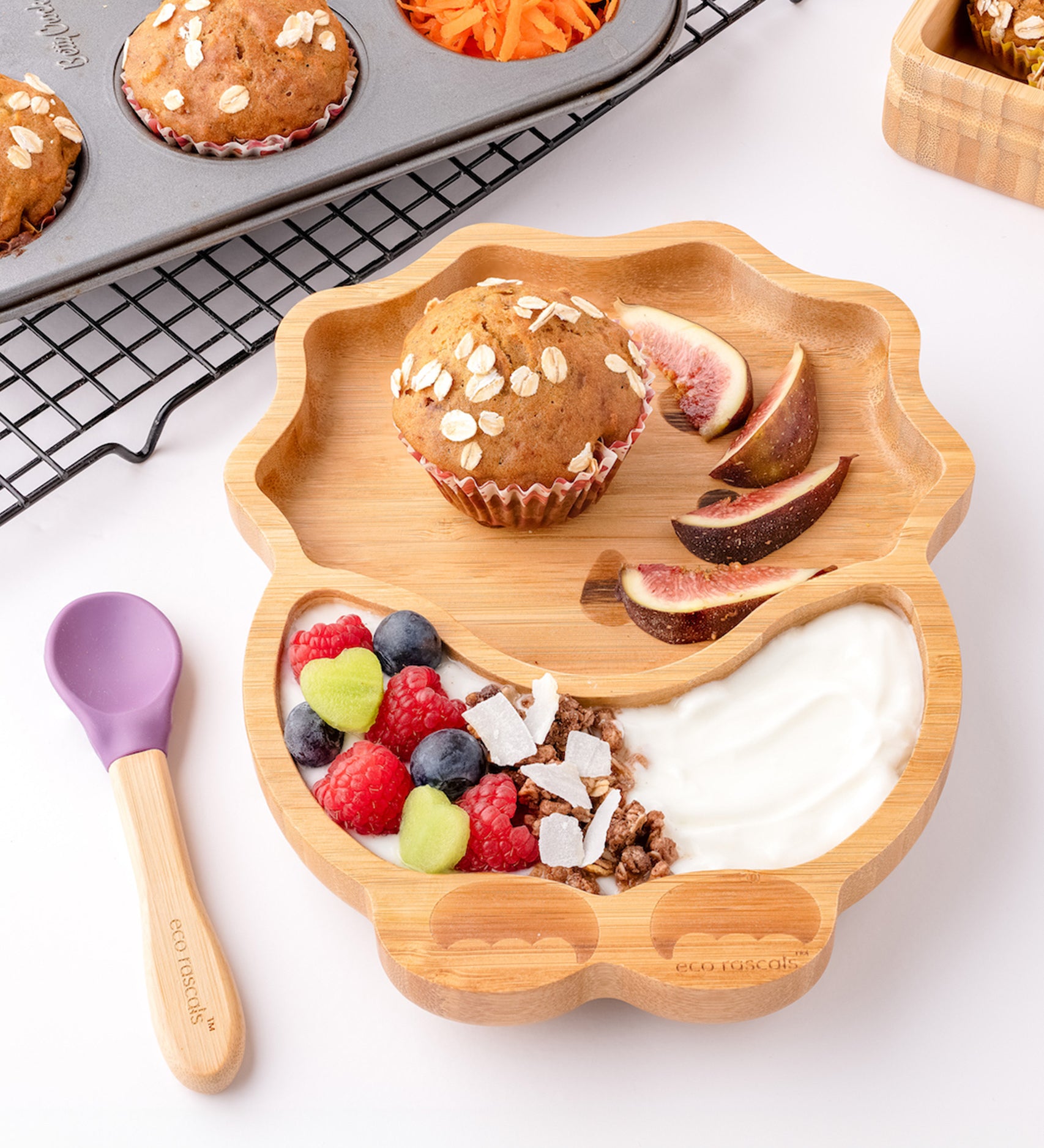 Eco Rascals Dinosaur Bamboo Suction Plate with a muffin, fig slices, yogurt and fresh fruit, and purple silicone and bamboo baby spoon.