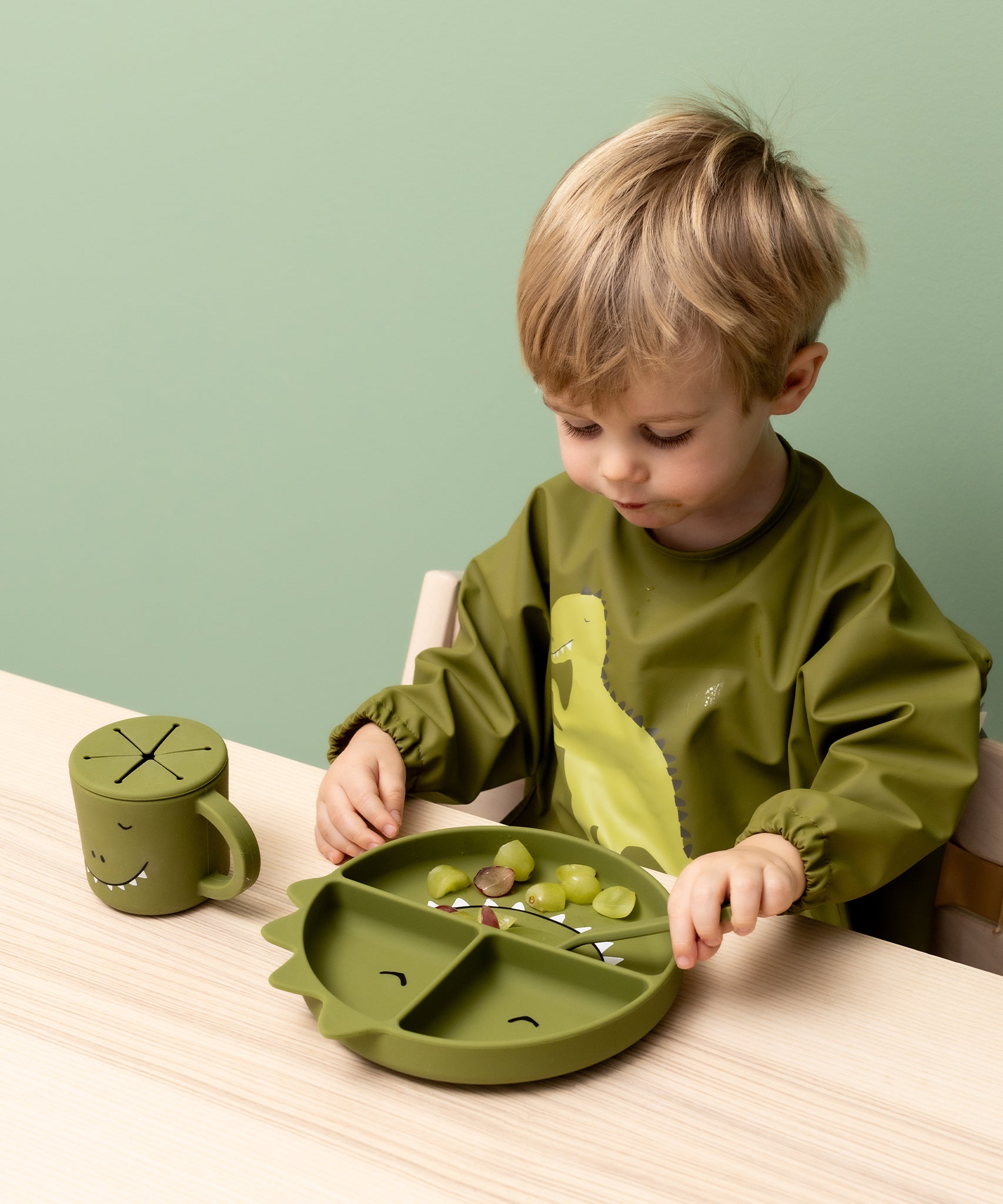 Child wearing the Trixie Animal Friends Waterproof Long Sleeve Bib, green, Mr Dino, with matching tableware.