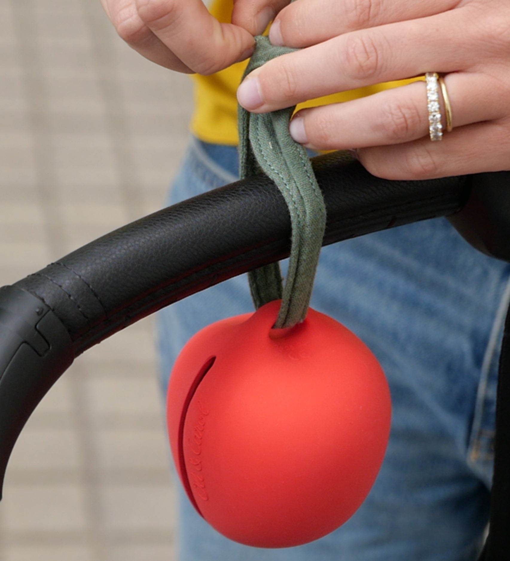 A person tying the Oli Carl Apple Pacifier Holder and Teether Toy made from natural rubber, onto the handle of their pram.