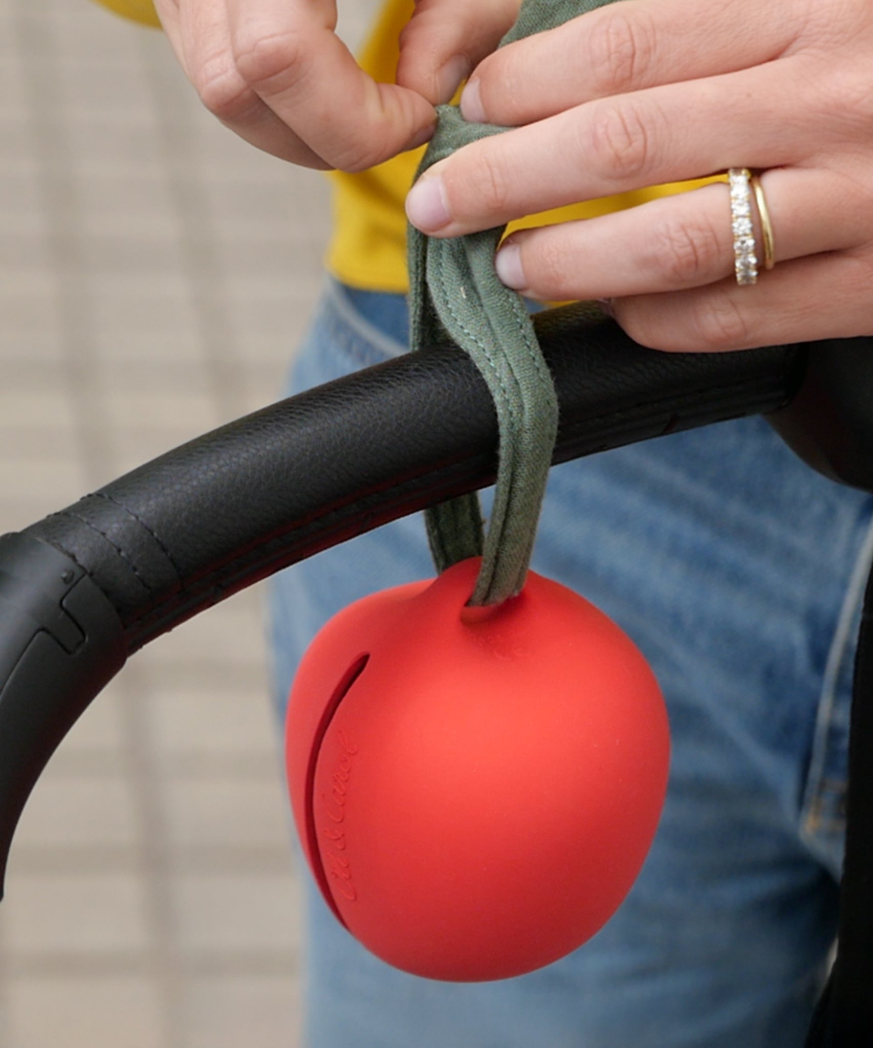 A person tying the Oli Carl Apple Pacifier Holder and Teether Toy made from natural rubber, onto the handle of their pram.