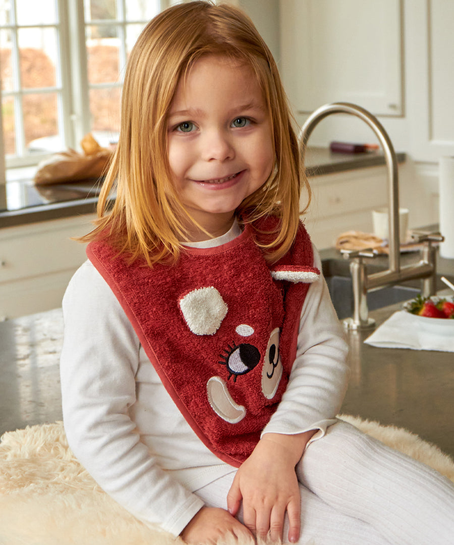 A child sat in a kitchen, wearing the Roommate Organic Cotton Baby Bib - Red Panda