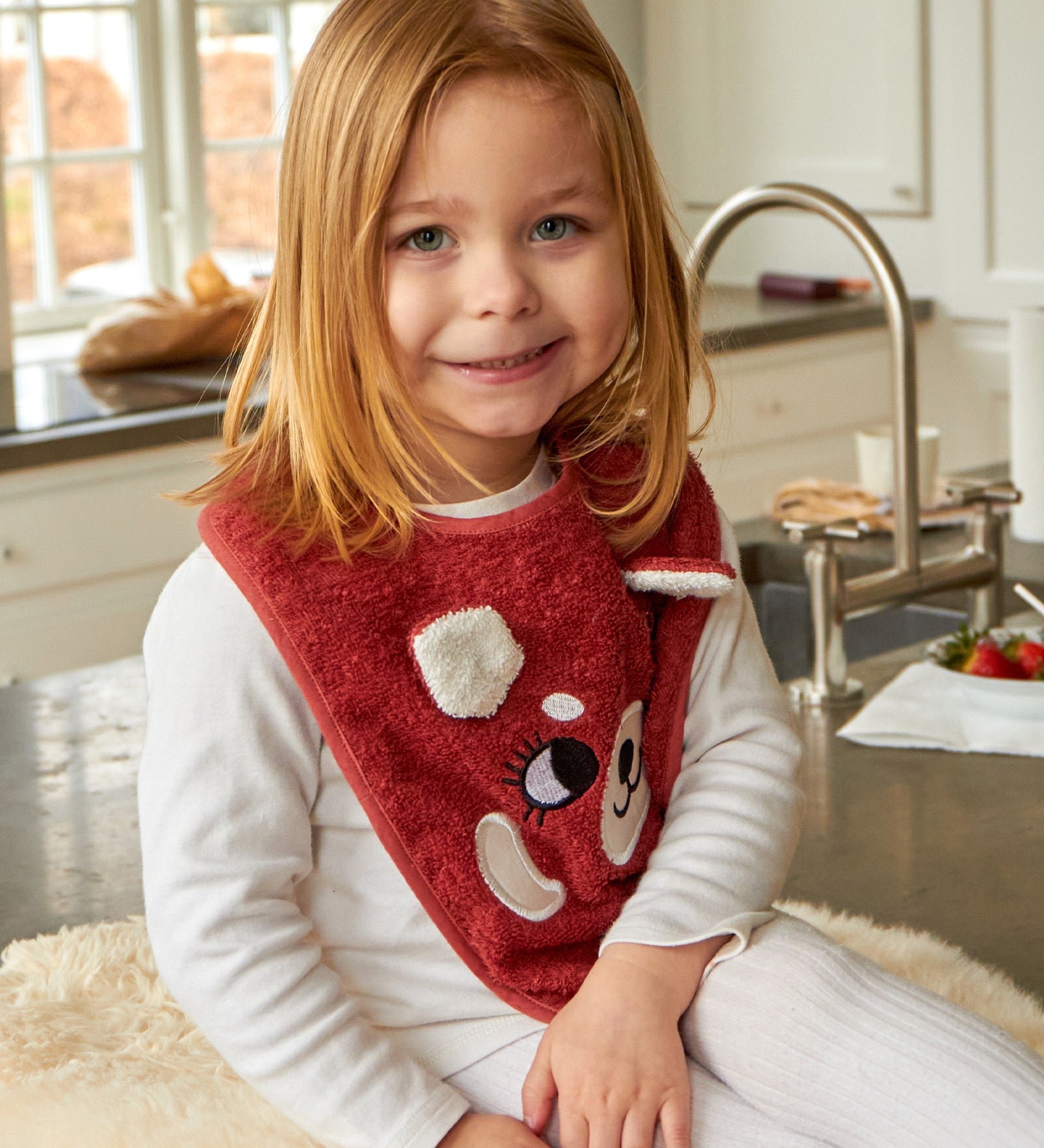 A child sat in a kitchen, wearing the Roommate Organic Cotton Baby Bib - Red Panda