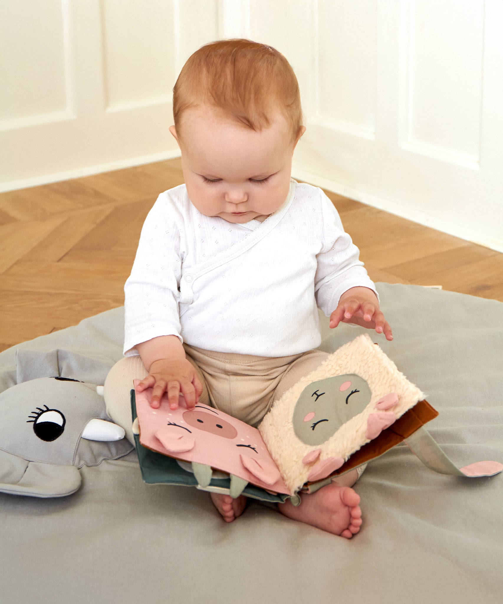 A baby sat on a Roommate playmat, playing with the Roommate Baby Nursery Book - Country Life on its lap. The fabric pages show a pig and a sheep