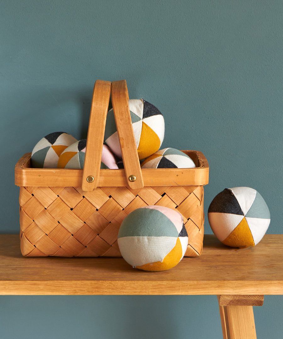 A collection of children's Roommate Canvas Balls With Bell stood on a wooden table in a wooden basket, with a blue/green background