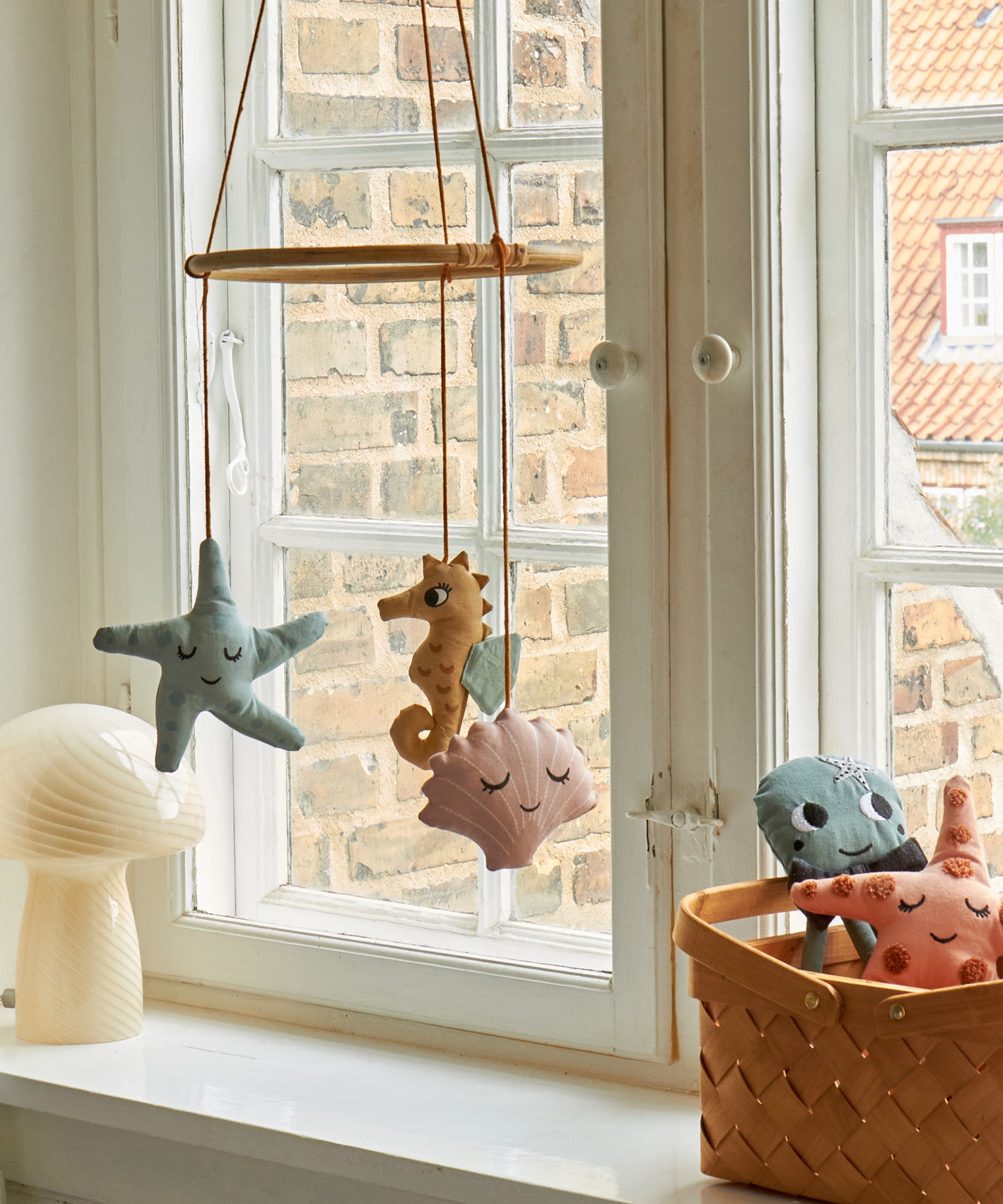 The Roommate Sealife Hanging Mobile hung in front of a window with Roommate toys in a basket and a mushroom lamp on the window ledge