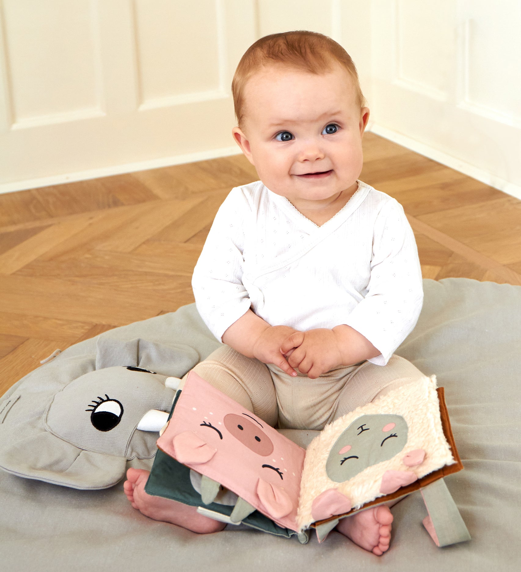 A baby sat on a Roommate playmat, with the Roommate Baby Nursery Book - Country Life on its lap. The fabric pages show a pig and a sheep