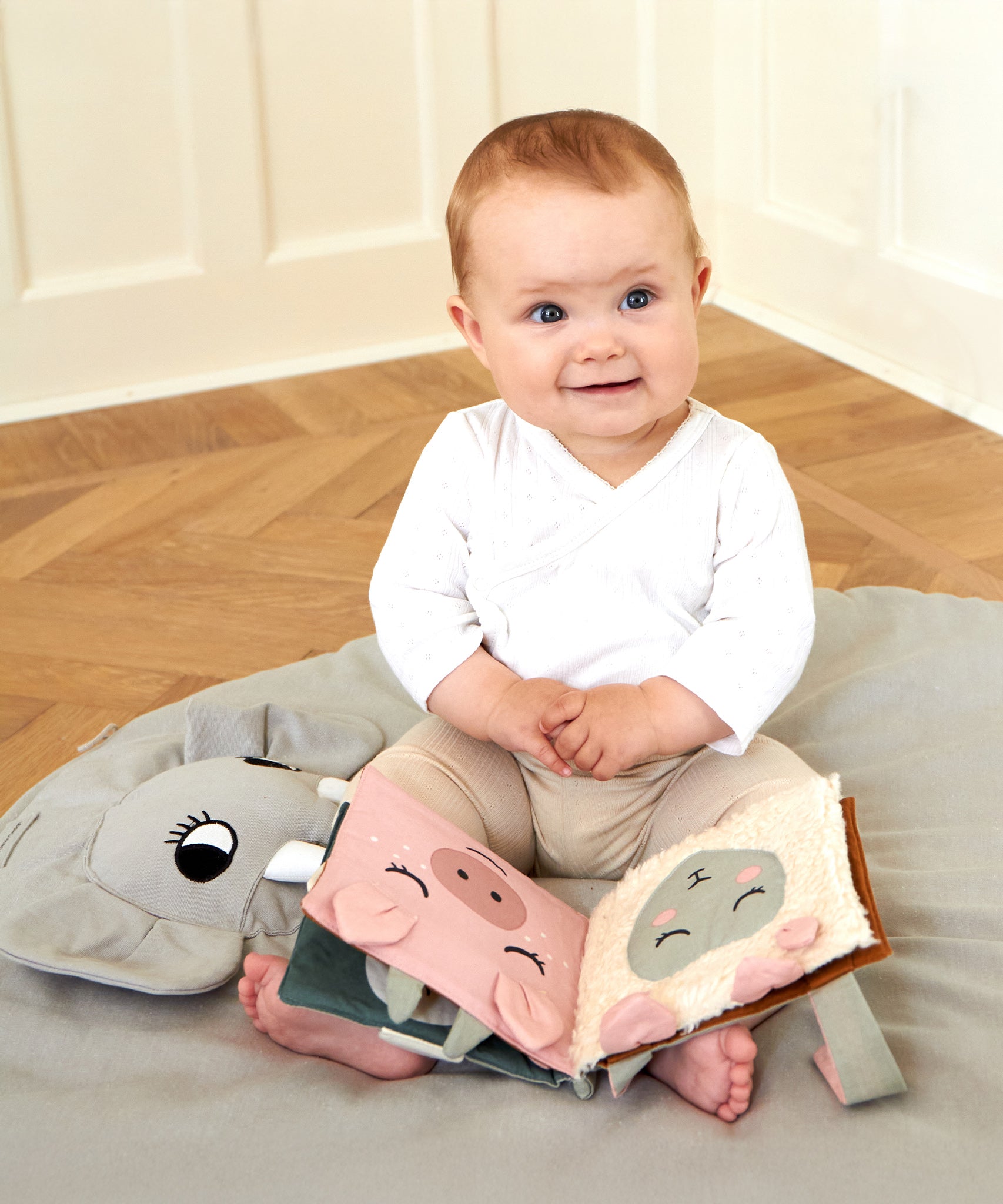 A baby sat on a Roommate playmat, with the Roommate Baby Nursery Book - Country Life on its lap. The fabric pages show a pig and a sheep