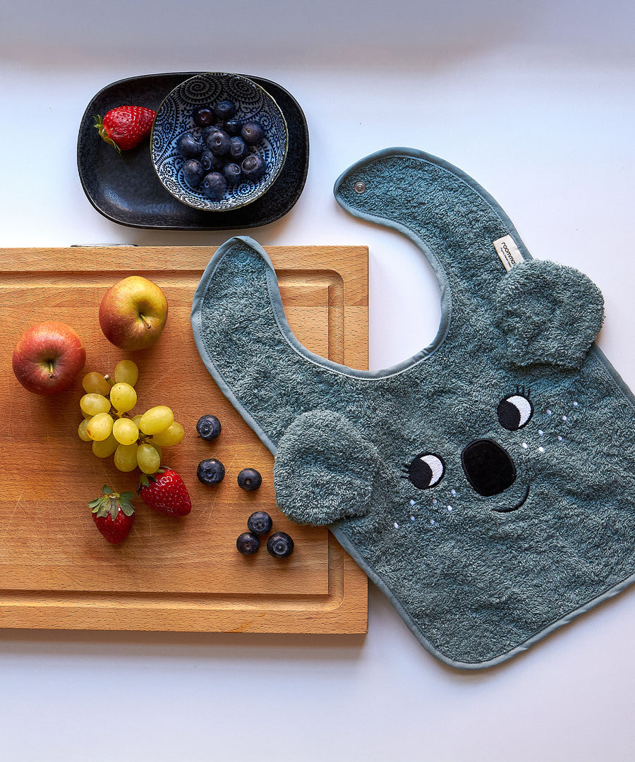 A top down view of the Roommate Organic Cotton Baby Bib - Koala on a white table top, half placed on a wooden chopping board with fruit on it. Above the chopping board is a black bowl with fruit inside