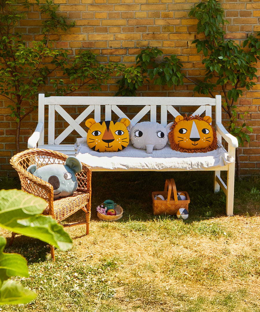 A collection of Roommate cushions, tiger, elephant, lion and koala on an outdoor garden bench in white, with a basket of Roommate Canvas balls underneath the bench