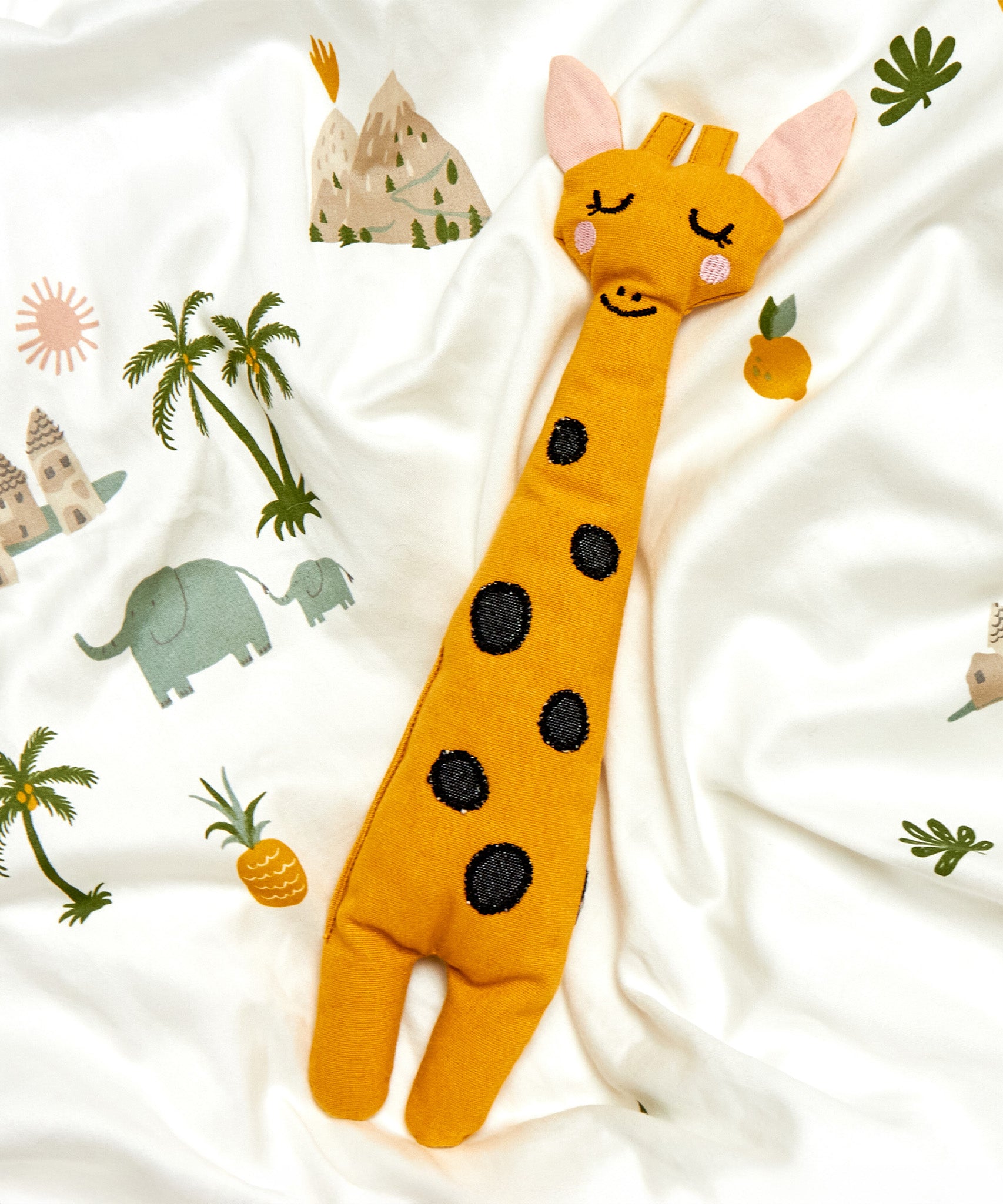 The Roommate Giraffe Rag Doll is laid on a white Roommate bed cover with printed animal and scenery pattern