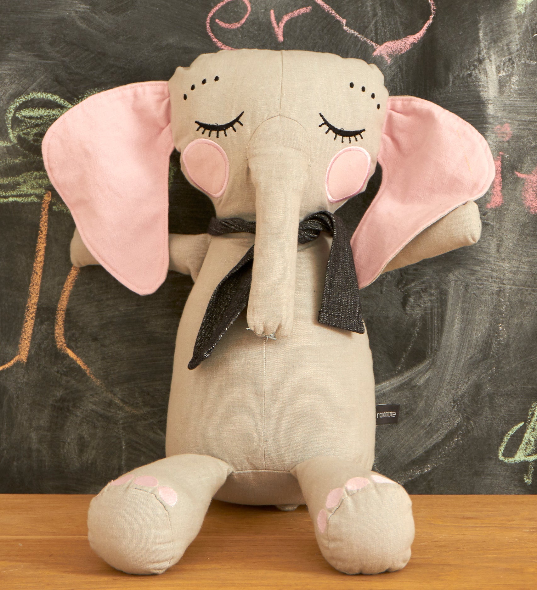 Roommate Little Grey Stuffed Elephant Toy sat on a wooden table with a chalkboard behind