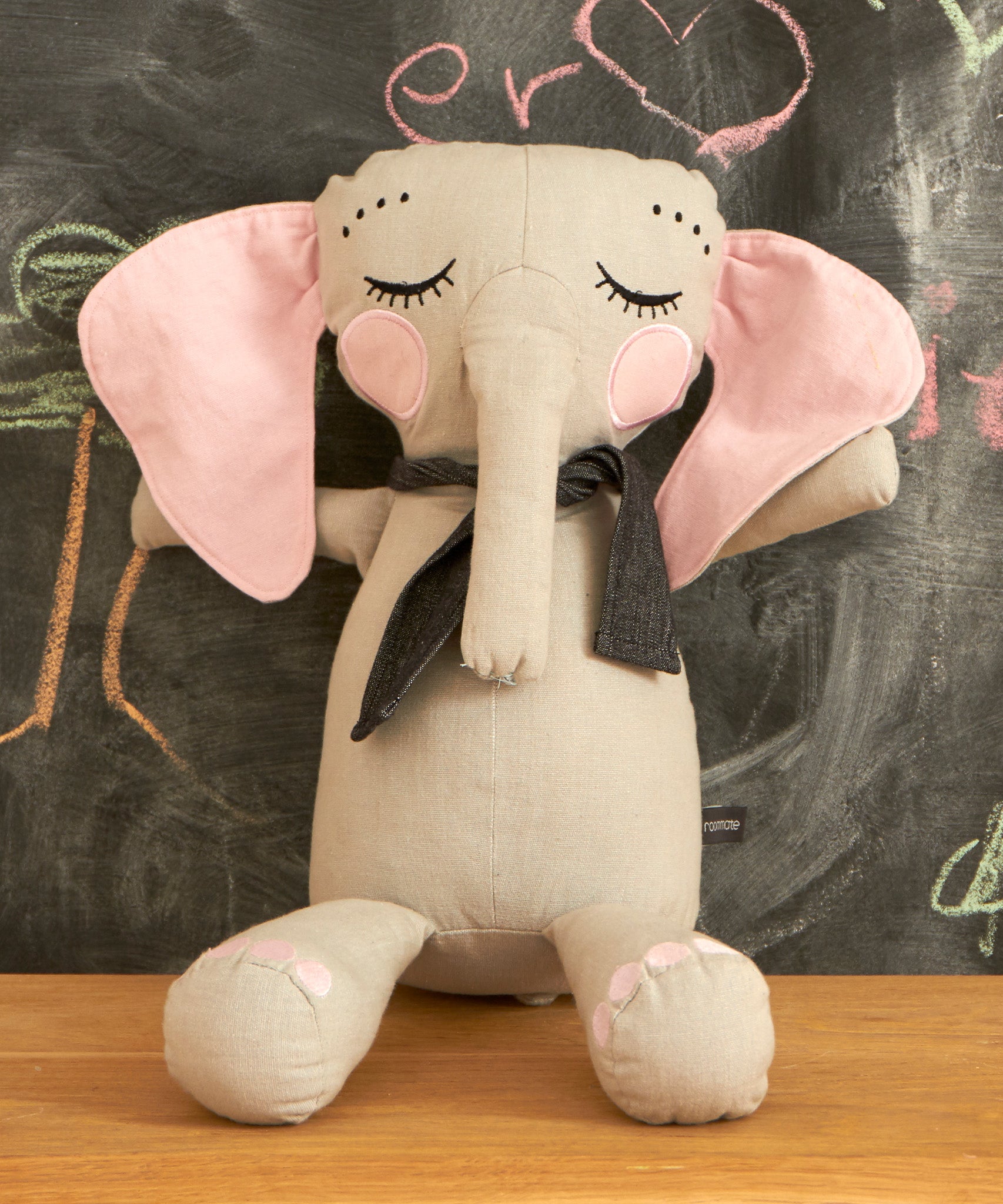 Roommate Little Grey Stuffed Elephant Toy sat on a wooden table with a chalkboard behind