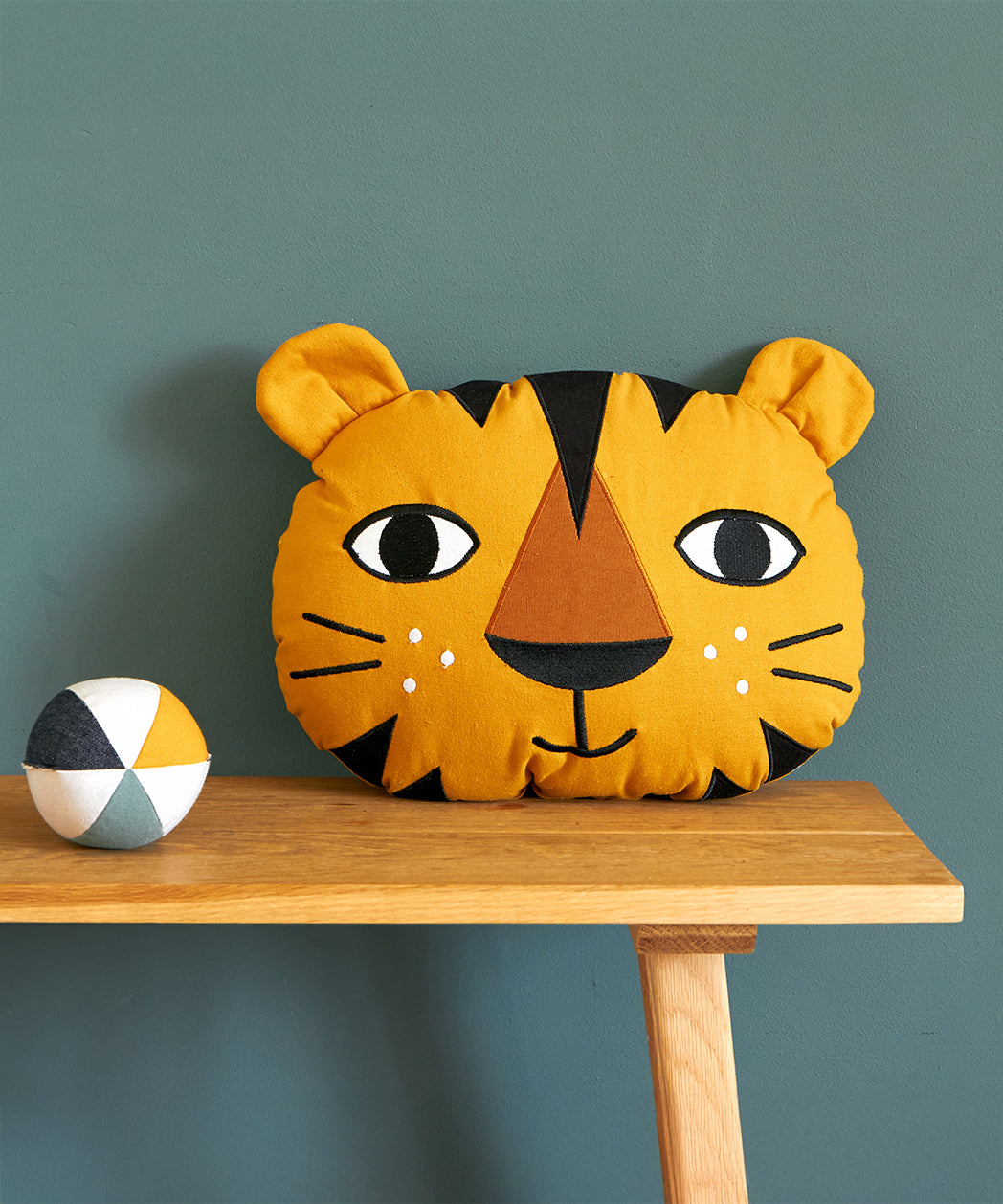 The Roommate Tiger Cushion is sat on a wooden table with a Roommate Canvas ball next to it. The image is on a dark teal background